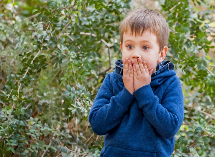 A blond, blue-eyed child covers his mouth with both hands in an expression of dismay