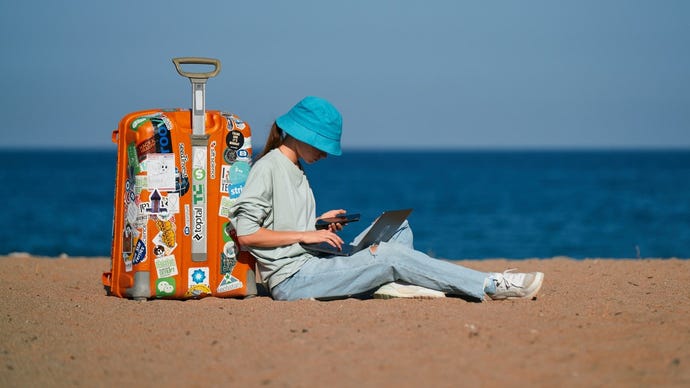 Woman in jeans and a blue hat holding a phone and laptop while sitting on a beach with a suitcase