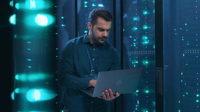 An analyst with a beard holding a laptop and standing in a server room.