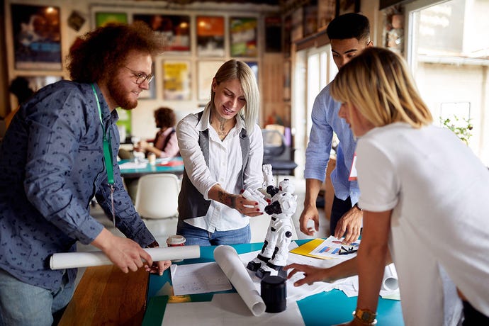 young adult creative people working together, standing around the desk with designs and a robot toy on it