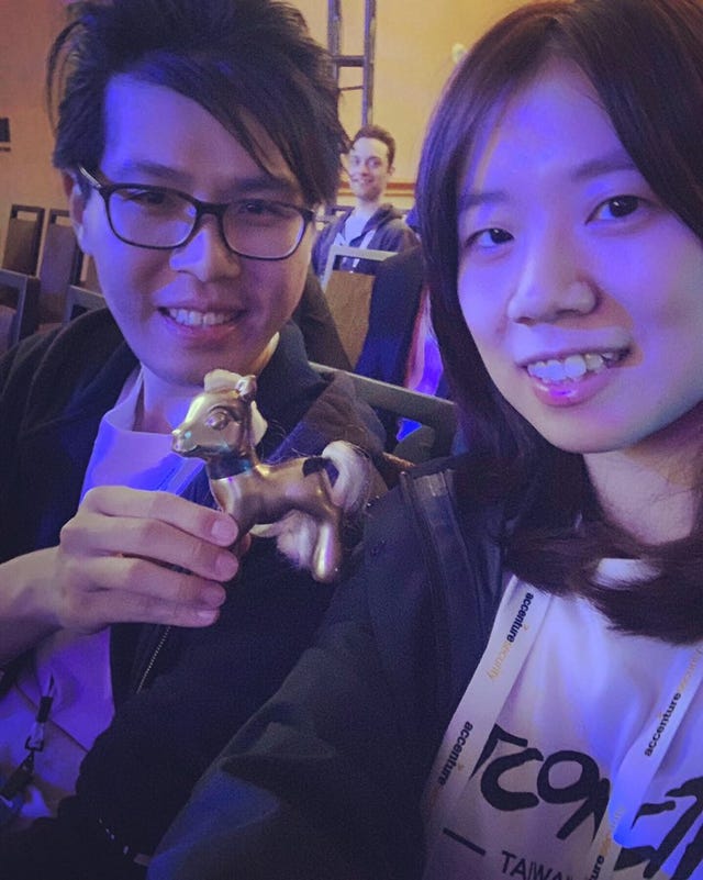 Two researchers, a man and a woman, take a selfie with their gold Pwnie statuette, photobombed by a smiling hoodie guy