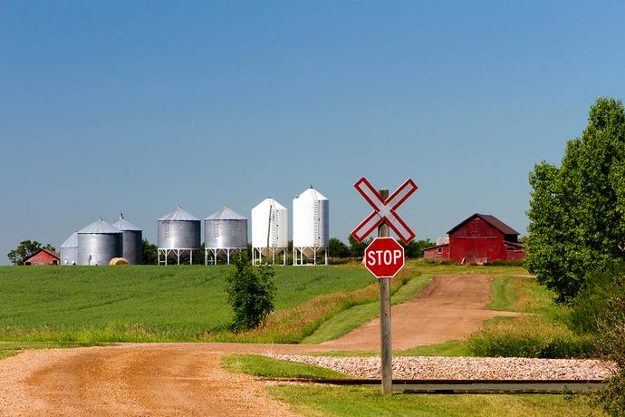 A railroad crossing with a stop sign in front of a farm with prominent siloes