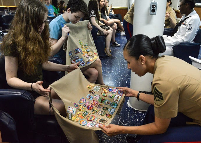 Two Girl Scouts hold up their patch-covered vests for a military woman to examine