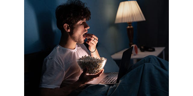 Young guy sits on the bed at night, enthusiastically watching movie on laptop and eating popcorn
