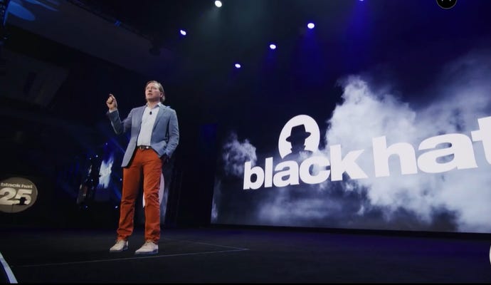 Chris Krebs stands on stage at Black Hat USA 2022. He's wearing a blue blazer, a button-down shirt, and orange pants.