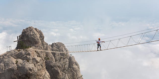 Man crossing a chasm on a hanging bridge, surrounded by clouds