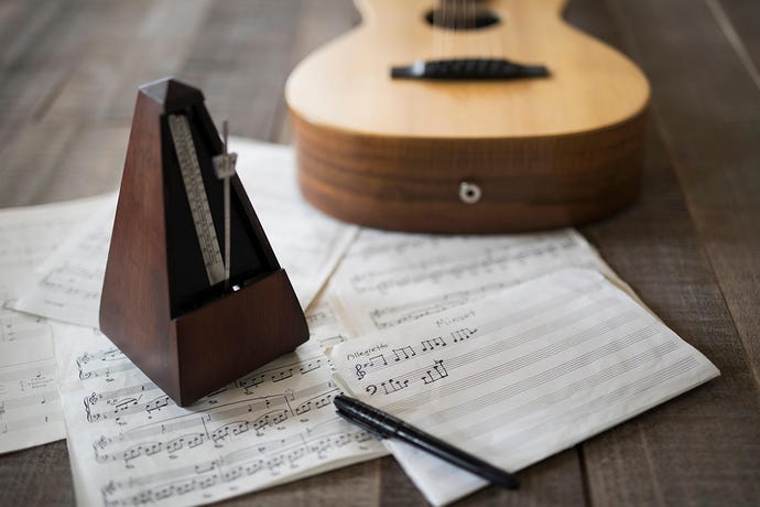 Photo of a wooden metronome on a pile of half-written sheet music with a guitar in the background
