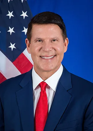 Keith Krach headshot as Under Secretary of State, in blue suit and red tie in front of an American flag