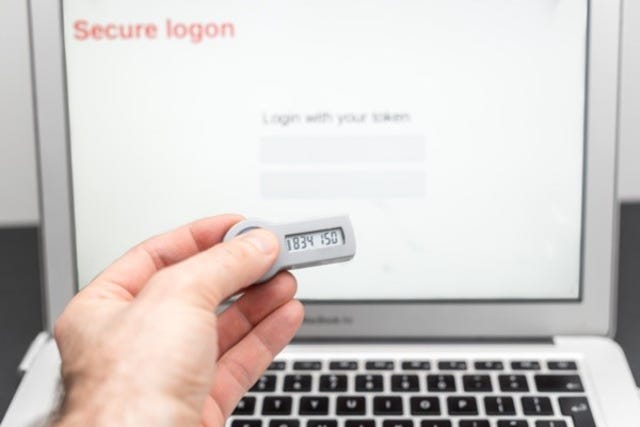 A white man's hand holds an authentication token keyfob with a generated access key in front of a monitor with Secure Logon