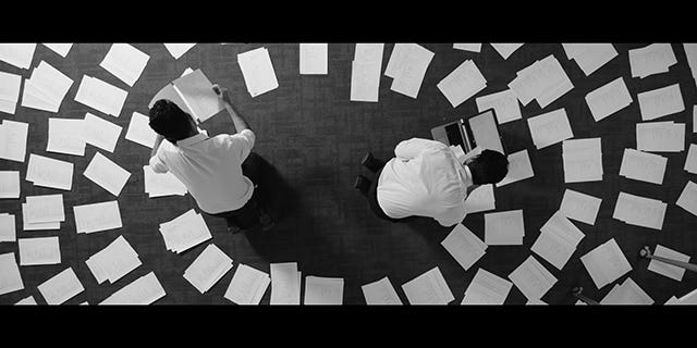 Two businessmen lay out hundreds of printouts in piles on the office floor