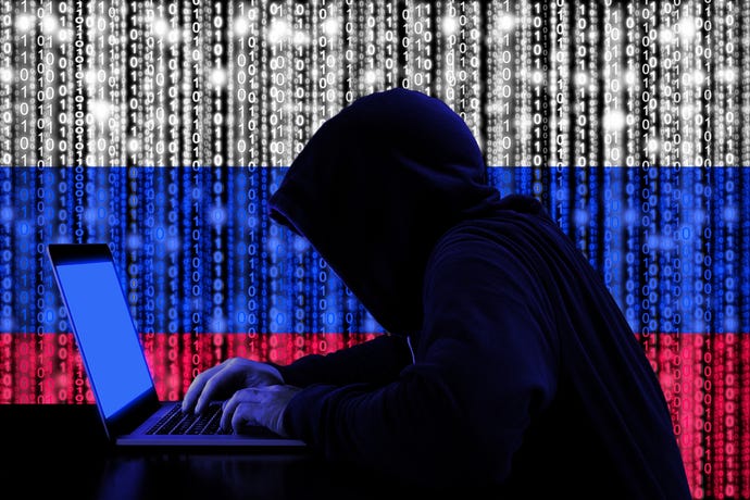 hooded figure hunched over a computer silhouetted against Russian flag