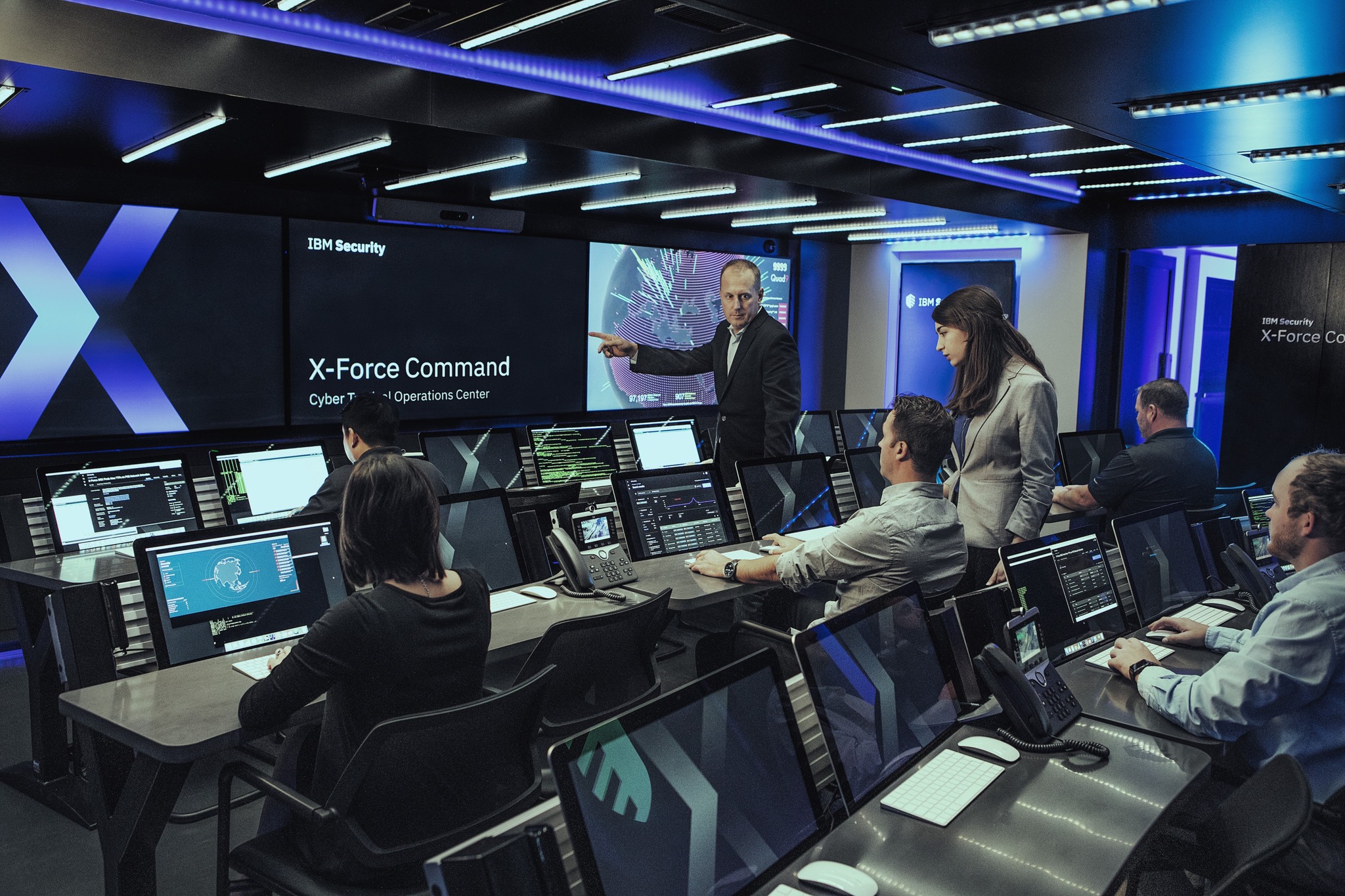  A group of cybersecurity analysts work at their computers in a dark room while a man and woman in business attire look at a large screen that displays a map of the world with various cyber threats highlighted.