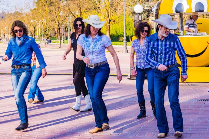 Line dancers wearing denim and cowboy boots move in sync in a public plaza
