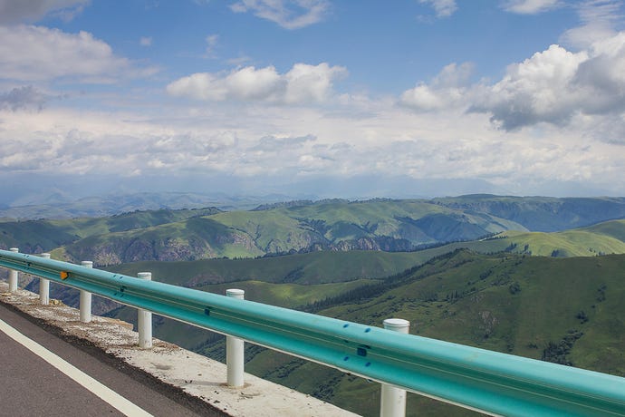 Photo of a green-painted guardrail lining a road on the edge of a dropoff in lush green mountains