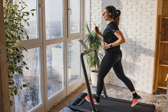 Image shows woman with a ponytail and headphones on jogging on a treadmill in front of a window in a home setting with plants