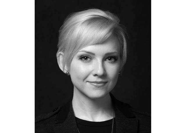 A blonde woman wearing a dark suit against a black background.