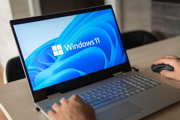 A man's hands type on a laptop whose screen displays the Windows 11 splash screen