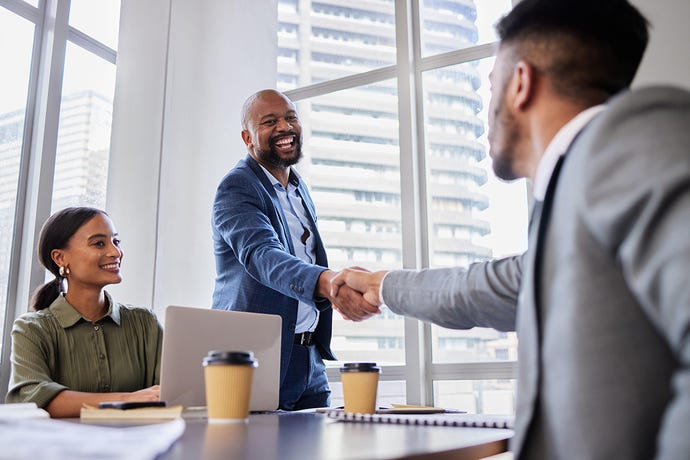 Shot of two business people shaking hands during a meeting while a third watches, typing on a laptop