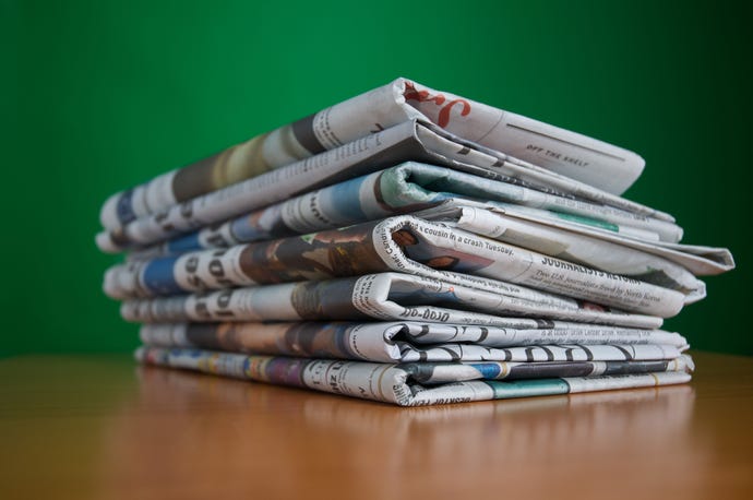 Image shows a stack of folded newspapers sitting on a light brown table against a green wall