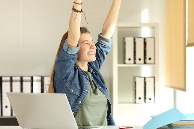 Young woman raises her arms exuberantly as she sits in front of a computer