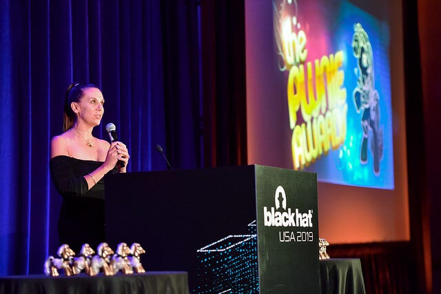 A woman in a black dress holds a microphone at a Black Hat USA 2019 lectern under a Pwnies banner