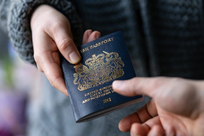 Closeup photo of hands: a white man in a blue sweater gives the new navy blue UK passport to another person