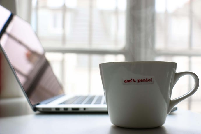 A teacup embossed with the phrase Don't Panic sits on a table next to an open laptop