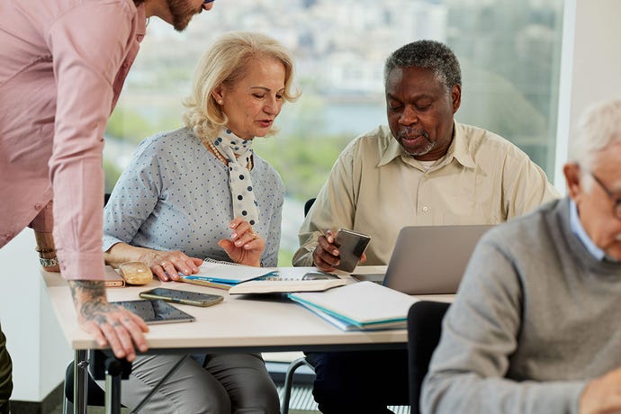 An older white woman and Black man learn and discuss the use of technology in a retraining course in the classroom.