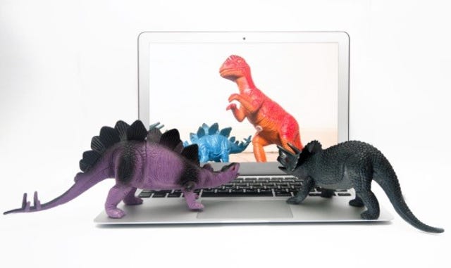 Toy stegosaurus and triceratops watch on a laptop as toy T. Rex attacks another stegosaurus