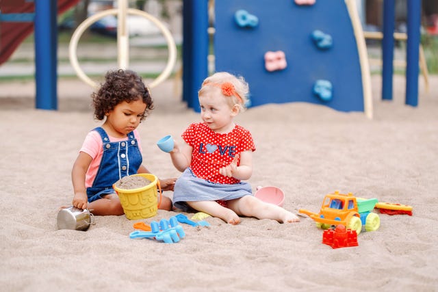 Two babies, one with light blonde hair and the other with dark curls, play together in a sandbox at a playground