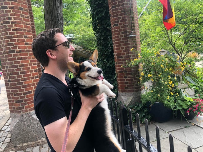 Zane Lackey, a man with short dark hair, wears sunglasses as he holds a corgi puppy on a front porch