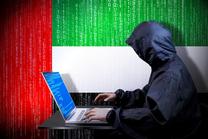 A hooded attacker sitting at a laptop in front of a UAE flag