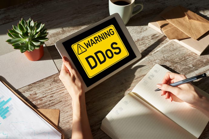 Image shows someone sitting at a table holding a tablet with a screen showing "Warning DDoS" in black writing in a yellow box