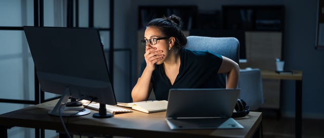 A woman looks horrified sitting in front of a computer, her hand is over her mouth in shock