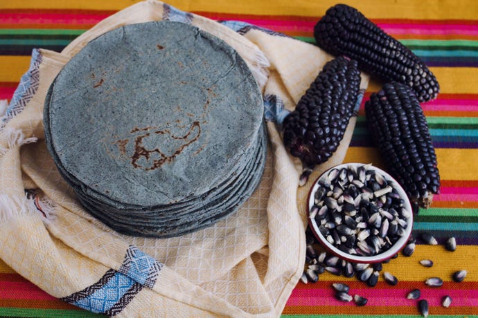 Stack of blue corn tortillas on a colorful cloth next to blue corn on cob and in kernel form