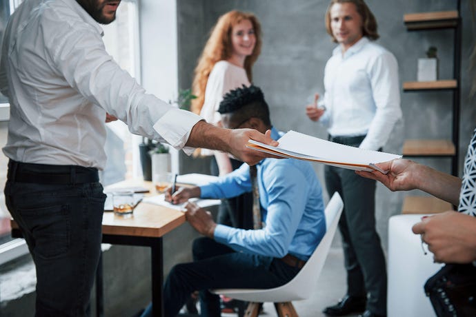 People in an office doing work, while in the foreground a man hands a folder to a business partner