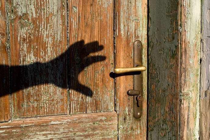 Photo of the shadow of a hand appearing to reach for the handle of an old wooden door