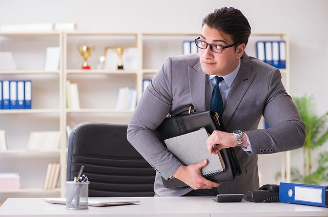 A person in a suit and black glasses in an office holding a bag and notebooks and having a very sneak expression on the face