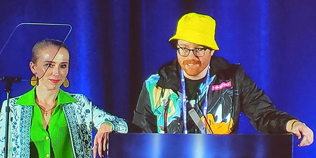 Sophia d'Antoine, a woman in a green dress, and Ian Roos, a man in a yellow bucket hat, at a Black Hat USA 2022 podium
