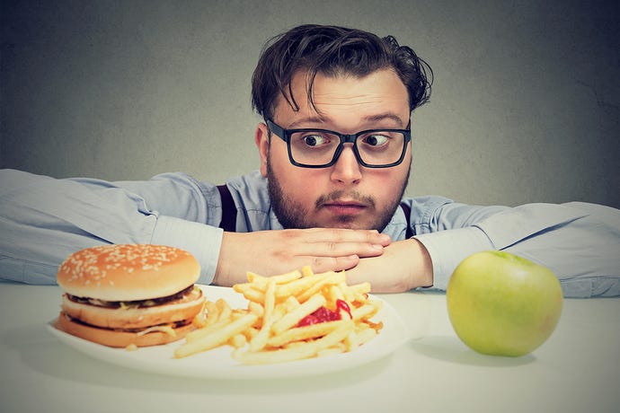 Young man with mustache and glasses leans down and stares anxiously at a burger and an apple