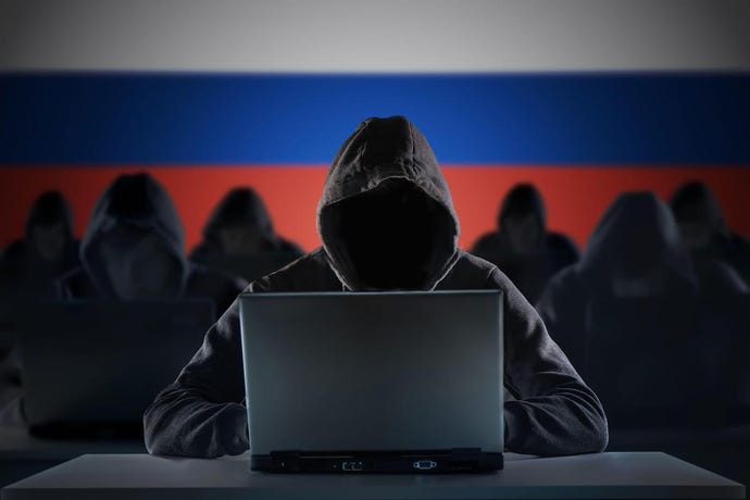 individuals wearing black hoodies behind their laptops and in front of a Russian flag.