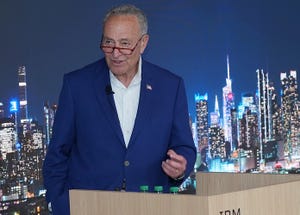 Senator Chuck Schumer speaking at a podium with a backdrop that depicts a cityscape.