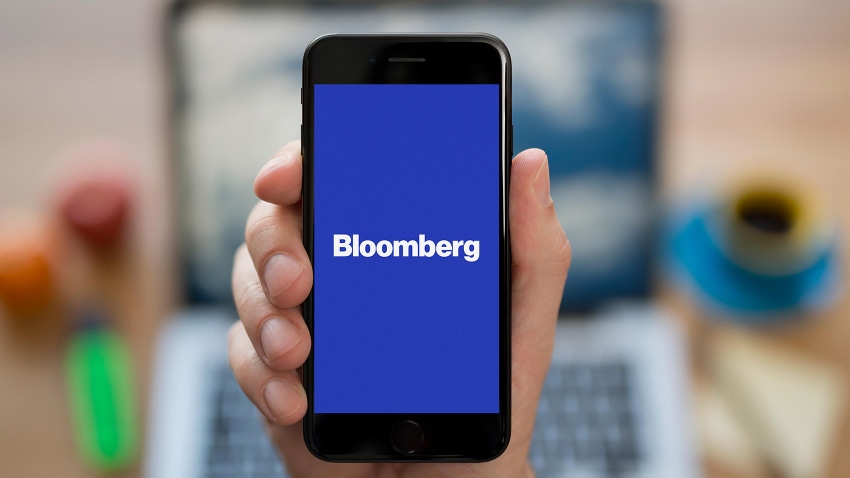 A man looks at his iPhone which displays the Bloomberg logo, while sitting at his computer desk 