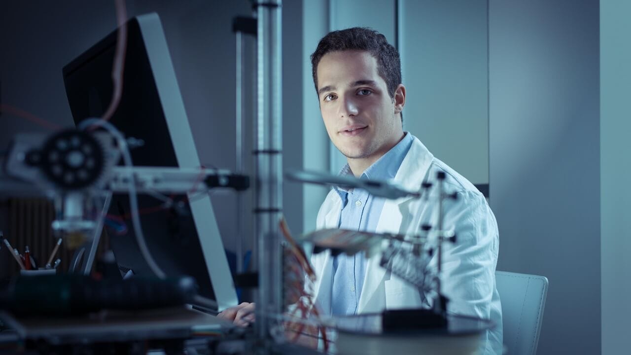 Researcher in the lab, wearing a lab coat and working with a computer, 3D printer on foreground. Researcher in the lab, wearing a lab coat and working with a computer, 3D printer on foreground.