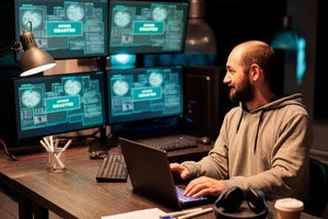 Young hacker stealing programming data from server, wearing hoodie with multiple screens.