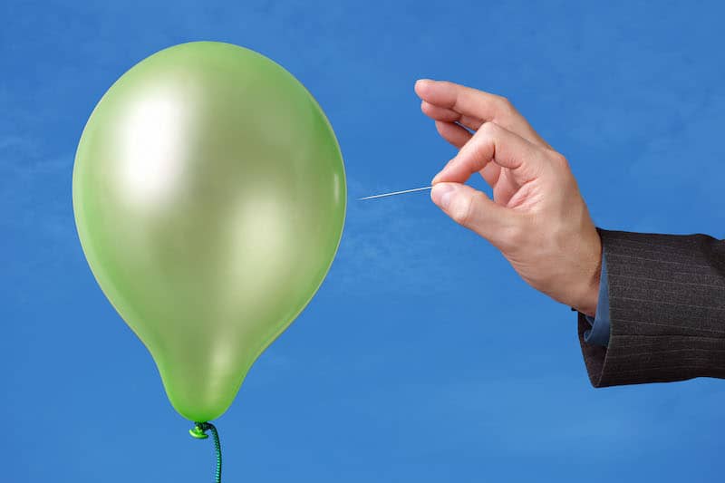 Green Balloon about to be burst by a needle held by a businessman's hand in a pinstripe sleeve.