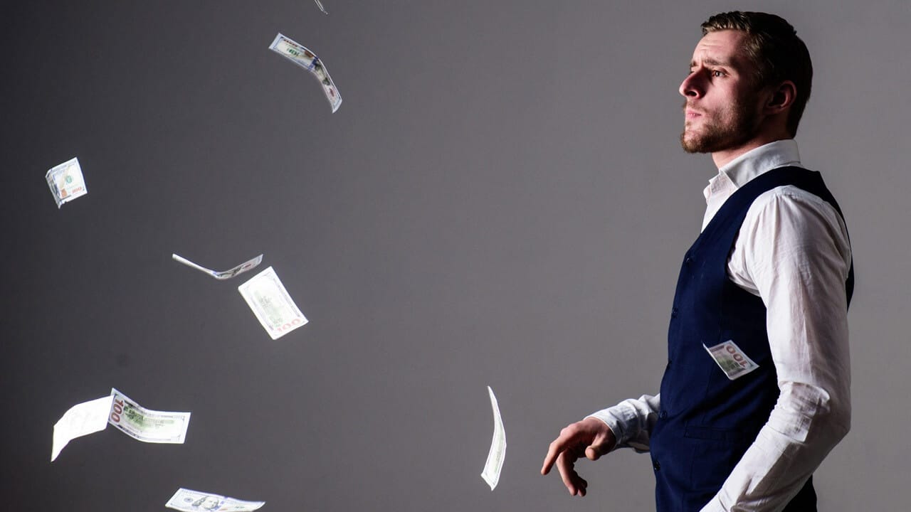 Man with strict face wasting money. Banknotes, cash dollars fly in air around. Man in formal wear, businessman throwing money on grey background. Man with strict face wasting money. Banknotes, cash dollars fly in air around. Man in formal wear, businessman throwing money on grey background.