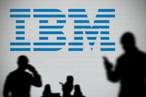The IBM logo is seen on an LED screen in the background while a silhouetted person uses a smartphone in the foreground.