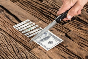 Close-up of hand cutting a 100-dollar bill with a sharp knife on a wooden table.
