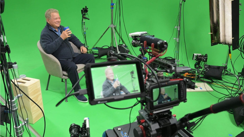 An old man sat in a chair on a green screen set surrounded by cameras